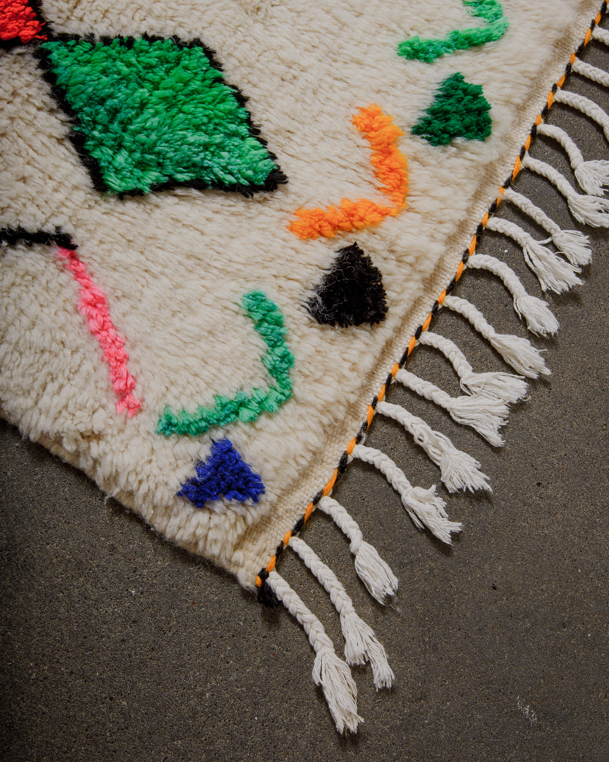 Corner of a Berberlin Finest rug with colorful patterns and tassels on a gray surface.
